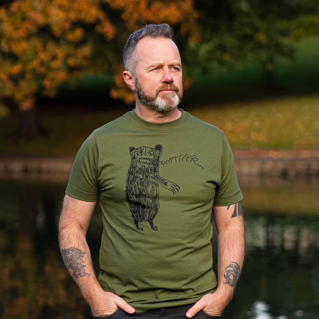 Man wearing an olive green graphic t-shirt with a growling bear design, standing outdoors in a park