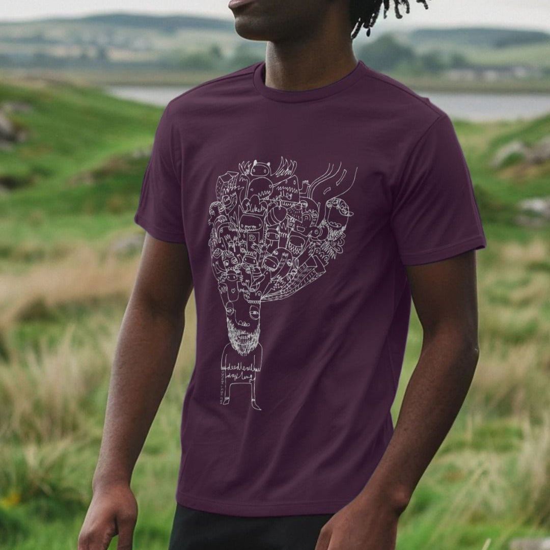 Man wearing a dark plum Doodleboy graphic t-shirt with an illustration of ideas bursting from a character's head, standing in a green landscape